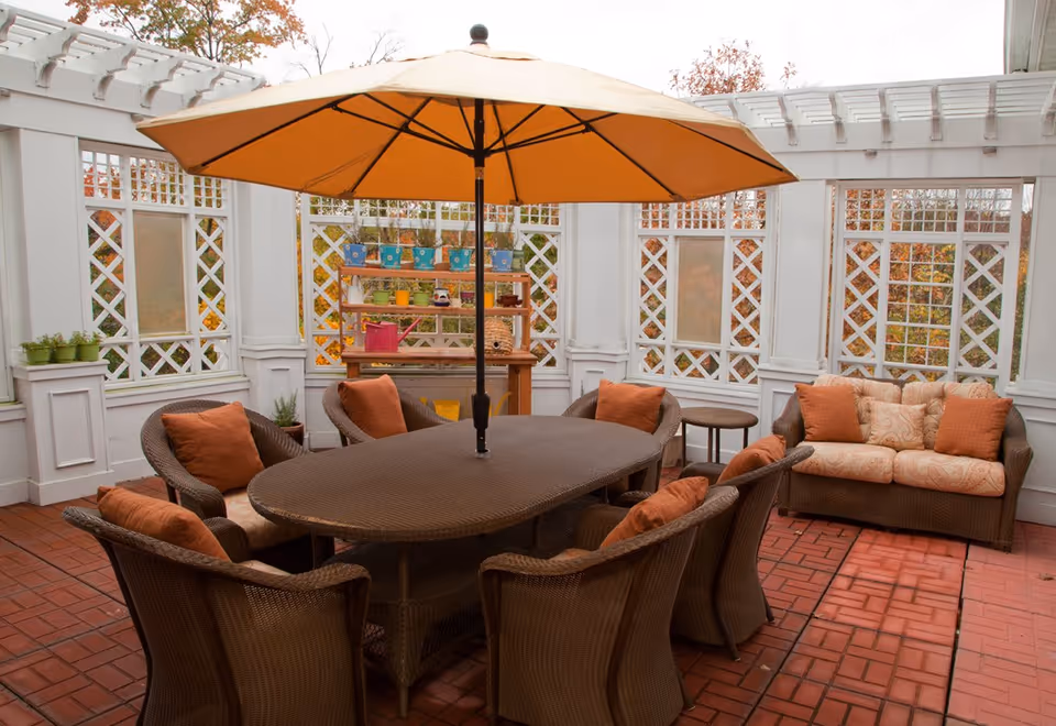 A cozy outdoor patio area with a large brown wicker table surrounded by six matching chairs with orange cushions. In the center of the table is a large beige umbrella. Behind the table is a wicker loveseat with patterned cushions and orange pillows. The patio is enclosed by white lattice walls with frosted glass panels and decorated with potted plants and gardening supplies on a wooden shelf.