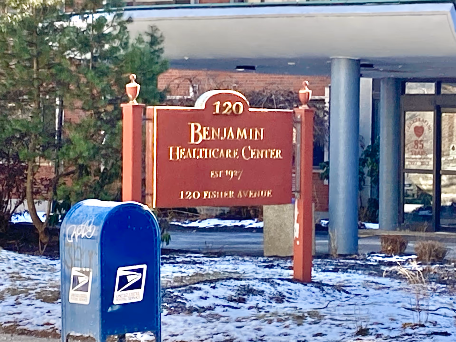 Outdoor view of the entrance area of Benjamin Healthcare Center with a red sign displaying the facility name and address, a blue USPS mailbox in the foreground, some snow on the ground, and trees and building columns in the background.