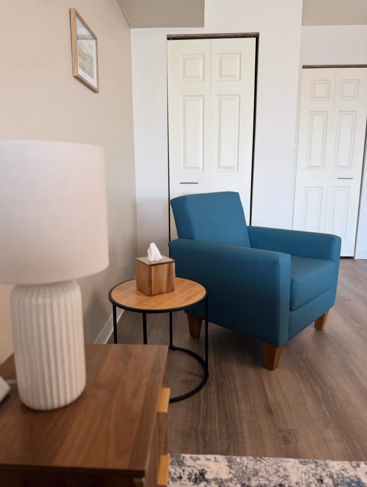 A cozy interior corner featuring a blue upholstered armchair next to a round wooden side table with a tissue box on it. In the foreground, there is a wooden surface with a white ribbed table lamp. The background shows white double-door closets and a beige wall with a small framed picture.