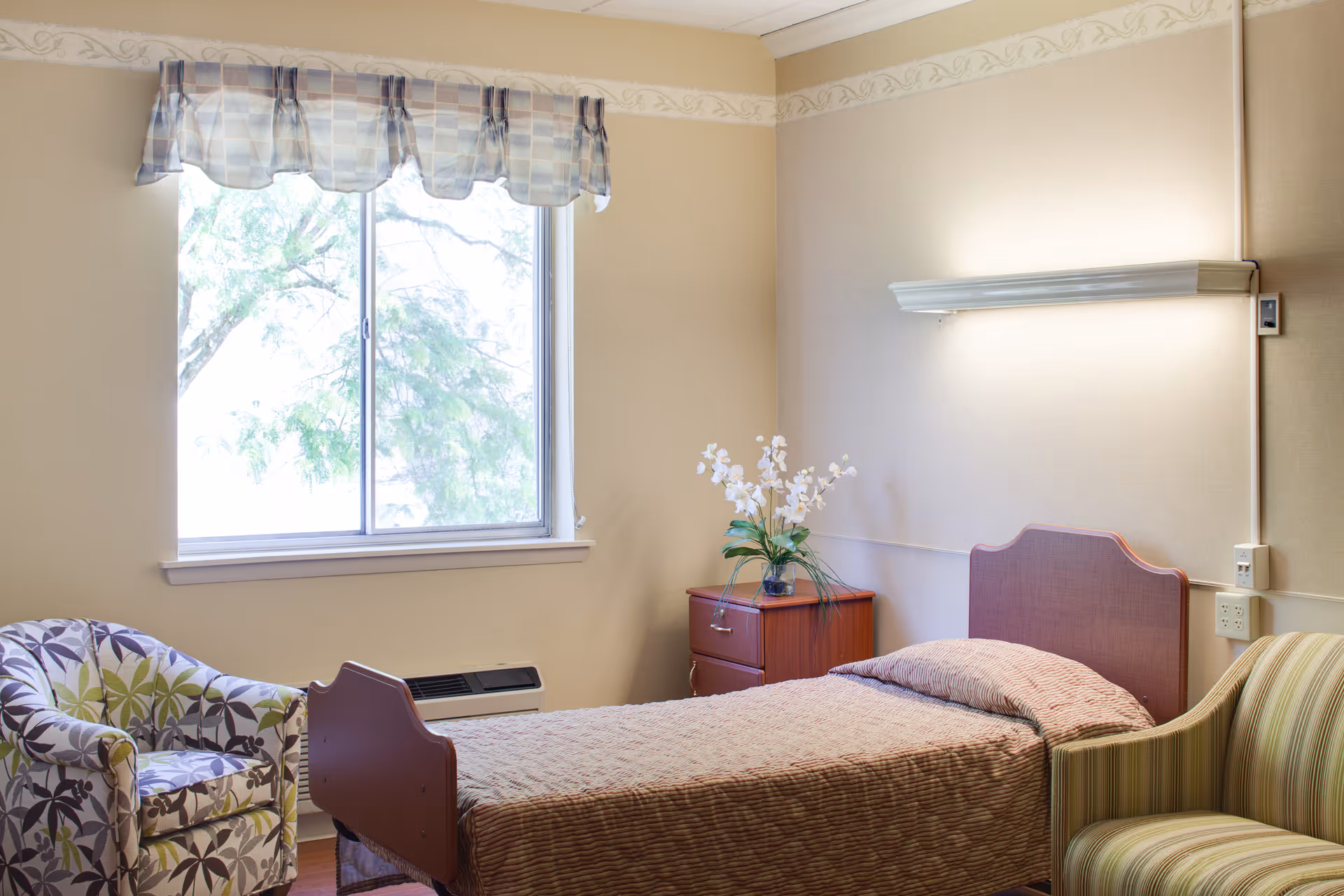 A tidy single-occupancy senior living bedroom with a bed, nightstand, window, and two chairs.