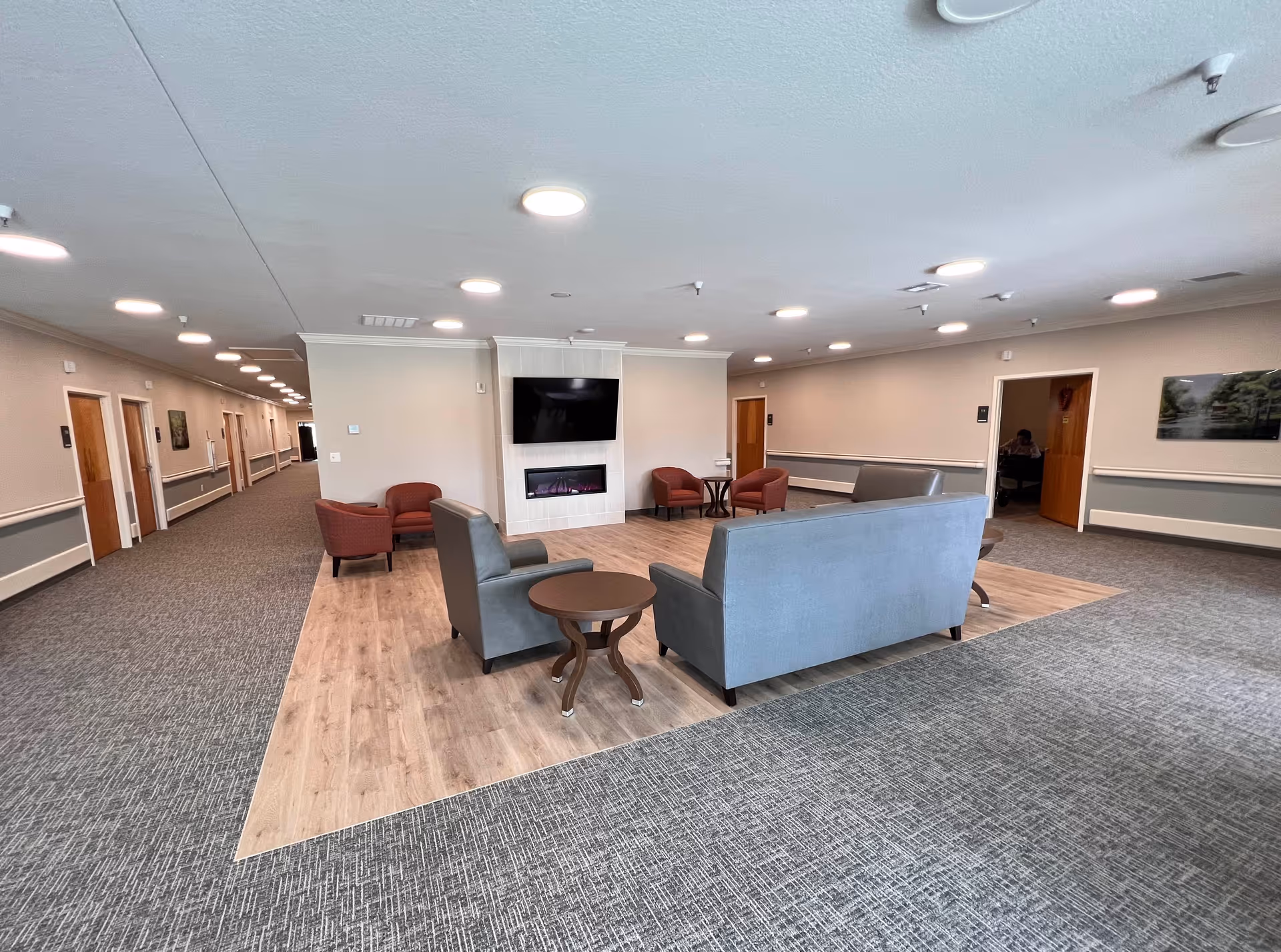 A spacious senior living facility common area with a seating arrangement including two gray sofas, two round wooden side tables, and four red armchairs. The seating area is centered on a wooden floor section surrounded by carpet. A flat-screen TV is mounted above a modern electric fireplace on the far wall. The room is well-lit with multiple ceiling lights and has beige walls with white trim. A long hallway with multiple doors extends to the left, and an open doorway on the right shows a person sitting inside another room.