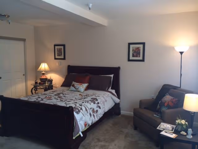A cozy bedroom featuring a dark wooden bed with floral bedding and multiple pillows. To the left of the bed is a small round table with a lamp and decorative items. On the right side of the room, there is a cushioned armchair with colorful pillows and a small side table holding a lamp, books, and a small flower arrangement. The walls are light-colored with two framed pictures hanging above the bed.