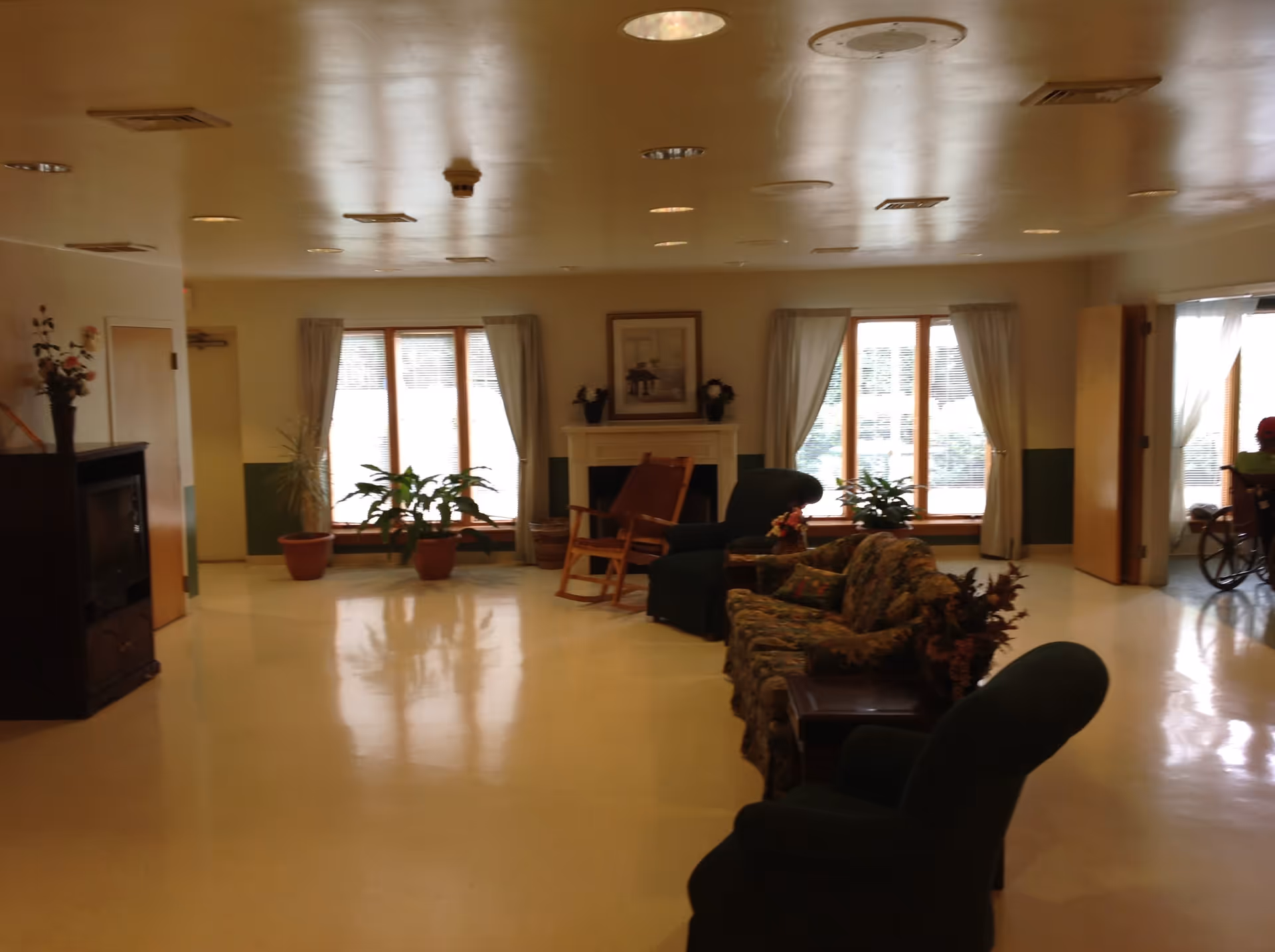 A spacious living room area in Mount Pleasant House with large windows covered by light curtains, a fireplace with framed artwork above it, several potted plants, a floral patterned sofa, a rocking chair, and multiple upholstered armchairs arranged around the room.