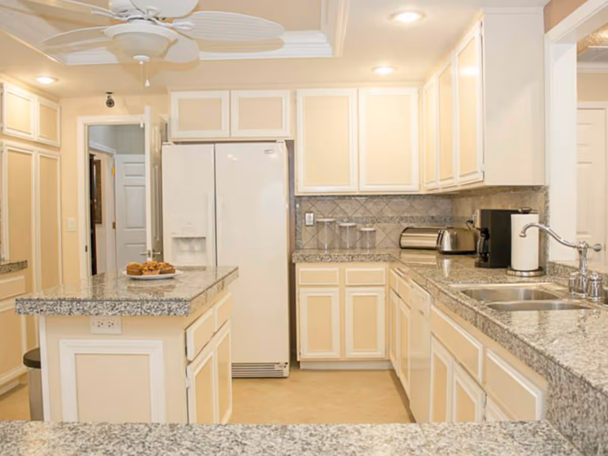 Bright kitchen with cream-colored cabinets and granite countertops. The kitchen features a white refrigerator, a double sink, a toaster, a coffee maker, and a paper towel holder. There is a ceiling fan with leaf-shaped blades and a small island with muffins on a plate.