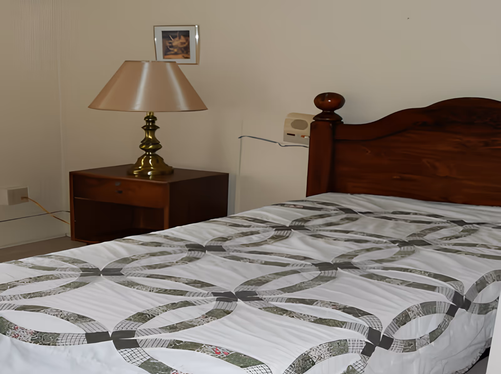 A bedroom with a wooden bed frame and a bed covered with a white quilt featuring a circular pattern in shades of gray and green. Next to the bed is a wooden nightstand with a brass lamp with a beige lampshade. A small framed picture hangs on the wall above the nightstand.