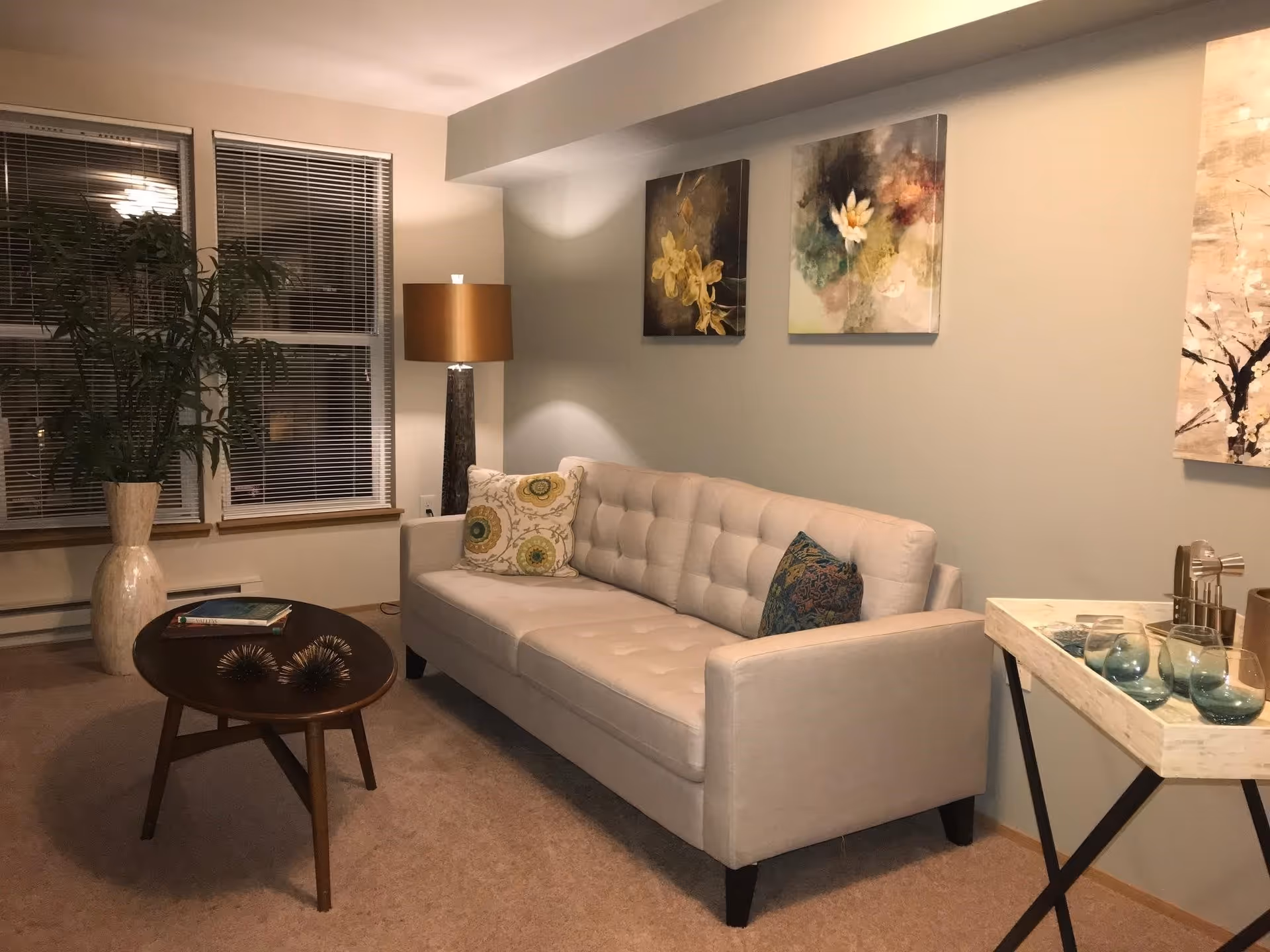 A cozy living room with a beige tufted sofa adorned with decorative pillows, a wooden coffee table with books and decorative items, a tall floor lamp with a brown shade, a large potted plant near two windows with blinds, and three floral-themed paintings on the wall. A small table with glassware is also visible on the right side.