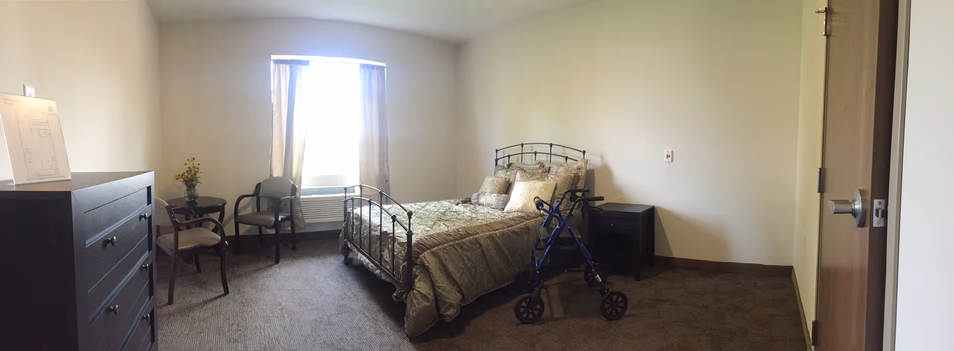 A senior living bedroom with a metal-framed bed covered in beige and gold bedding and multiple pillows. A blue walker is positioned next to the bed. There is a dark wooden dresser on the left side, two chairs and a small round table with a vase of yellow flowers near a window with beige curtains. A nightstand is next to the bed on the right side. The room has beige walls and carpeted floor.