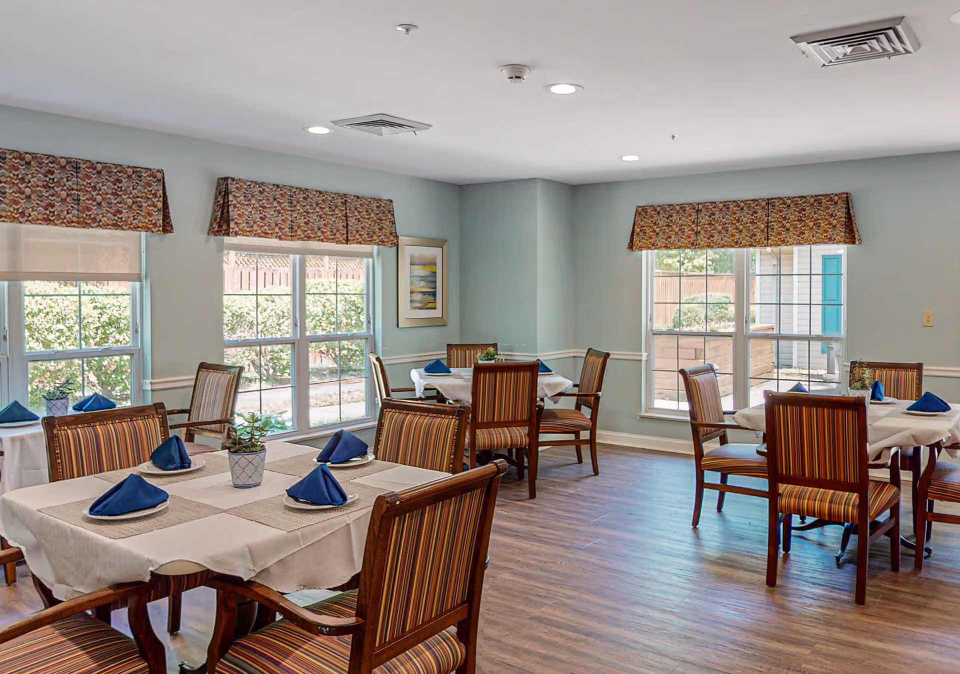 A dining room with several tables covered in white tablecloths and beige placemats, each set with plates and blue folded napkins. The chairs have wooden frames with striped upholstery. Large windows with patterned valances let in natural light, and there is a small potted plant on one of the tables. The floor is wood, and the walls are painted light blue with white trim.