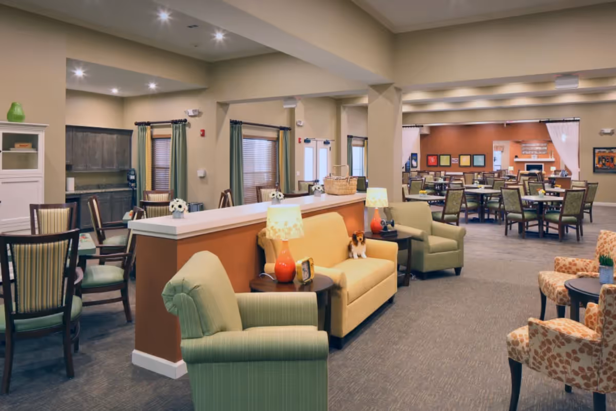 A spacious senior living facility common area with comfortable seating including green armchairs, a yellow sofa, and patterned chairs. There are side tables with lamps and decorative items. In the background, there are multiple dining tables and chairs arranged neatly, with framed artwork on the walls and a fireplace visible. The room has warm lighting and a welcoming atmosphere.
