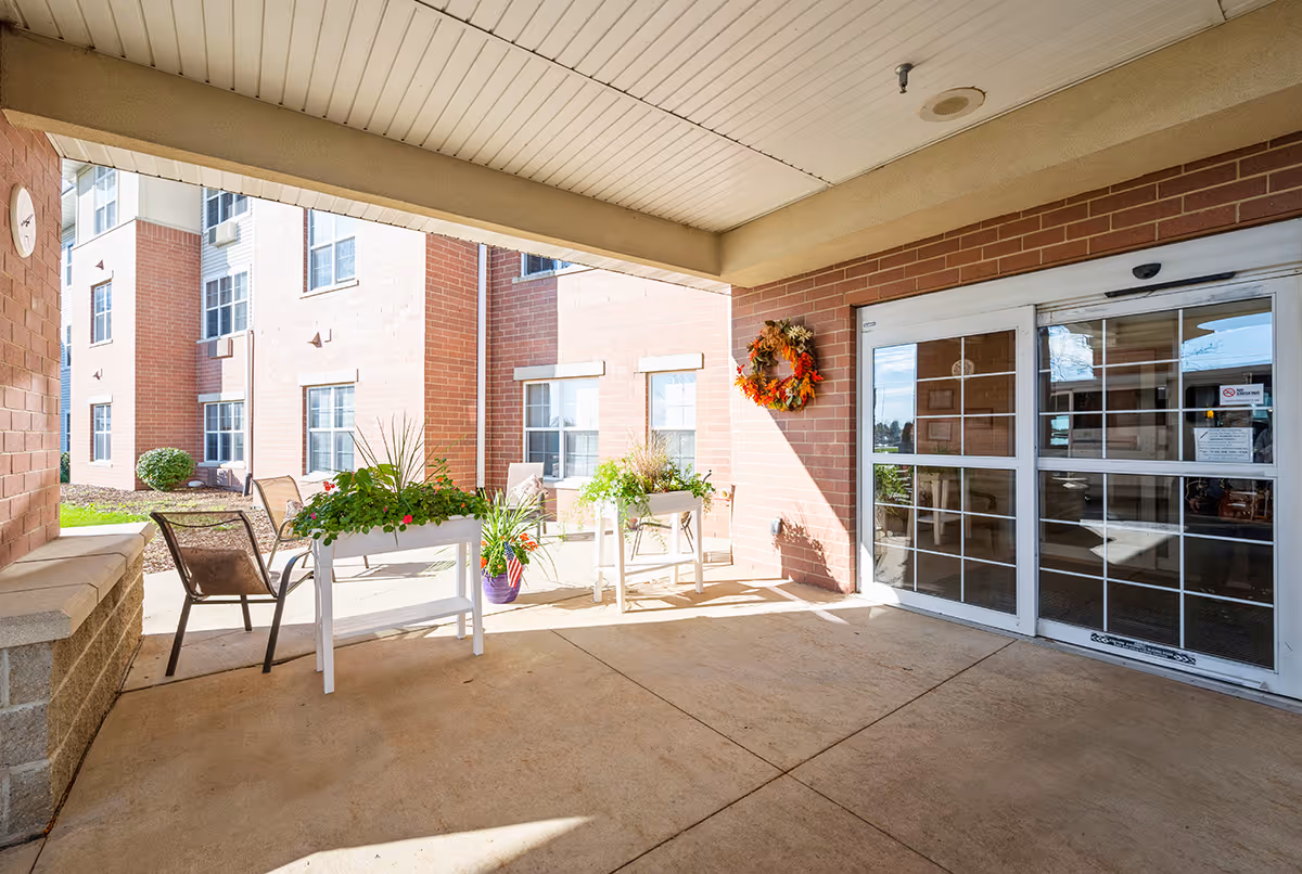 Covered entrance/porch of a senior living facility with chairs, potted plants, a wreath, and glass double doors.