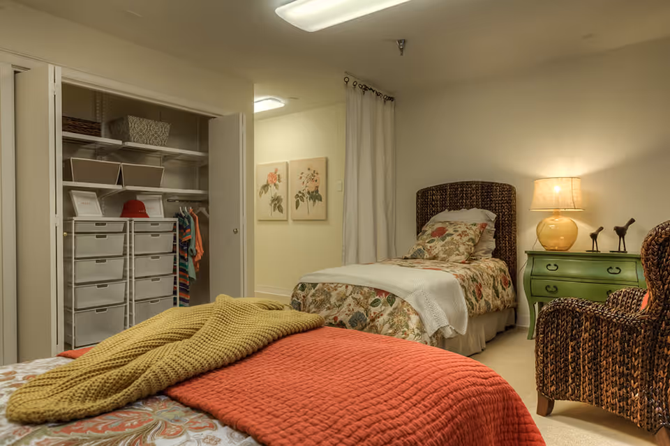 A cozy bedroom with two beds, one in the foreground with a red quilt and a yellow knitted blanket, and another in the background with floral bedding. There is an open closet with shelves and hanging clothes on the left side. A green nightstand with a lamp and decorative bird figurines is next to the bed in the background. A wicker chair is partially visible on the right side of the image. The walls are light-colored, and two floral paintings hang on the wall near the closet.