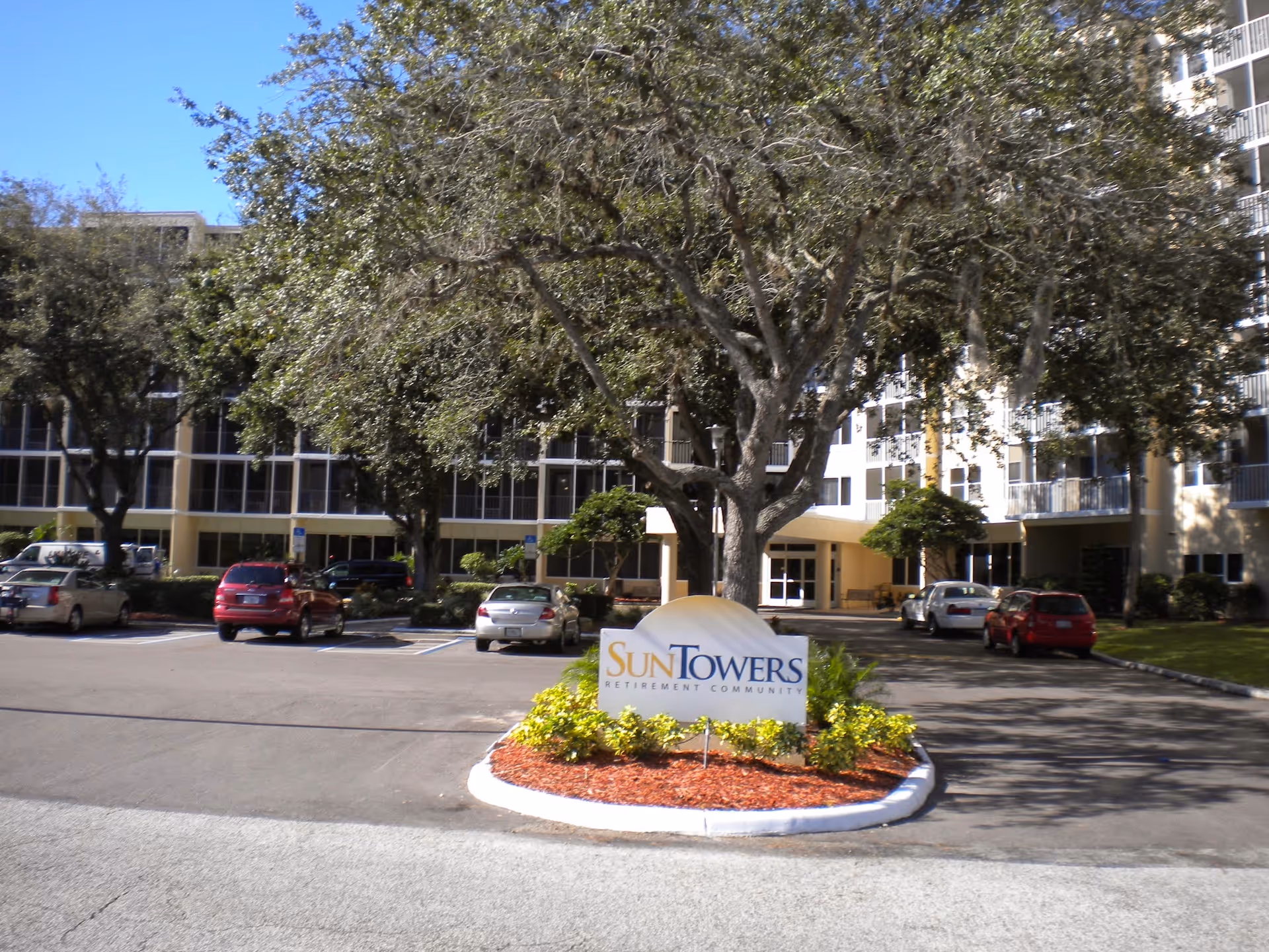 Front entrance of Sun Towers Retirement Community with a sign in a landscaped island, a large tree, and parked cars.