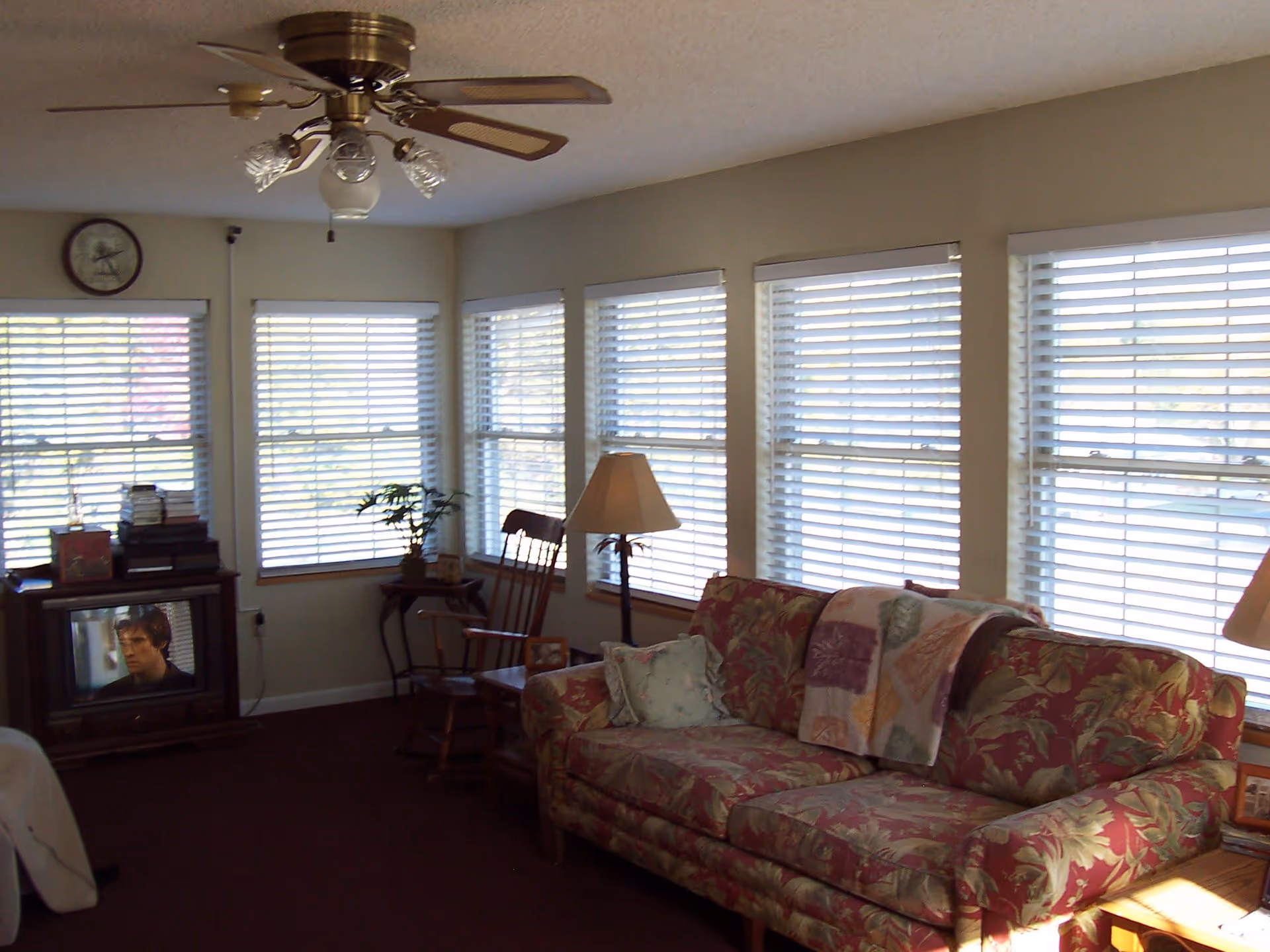 A cozy living room with a floral patterned sofa, a rocking chair, a side table with a lamp, and a small TV on a stand. The room has multiple windows with white blinds letting in natural light, and a ceiling fan with lights is mounted on the ceiling.