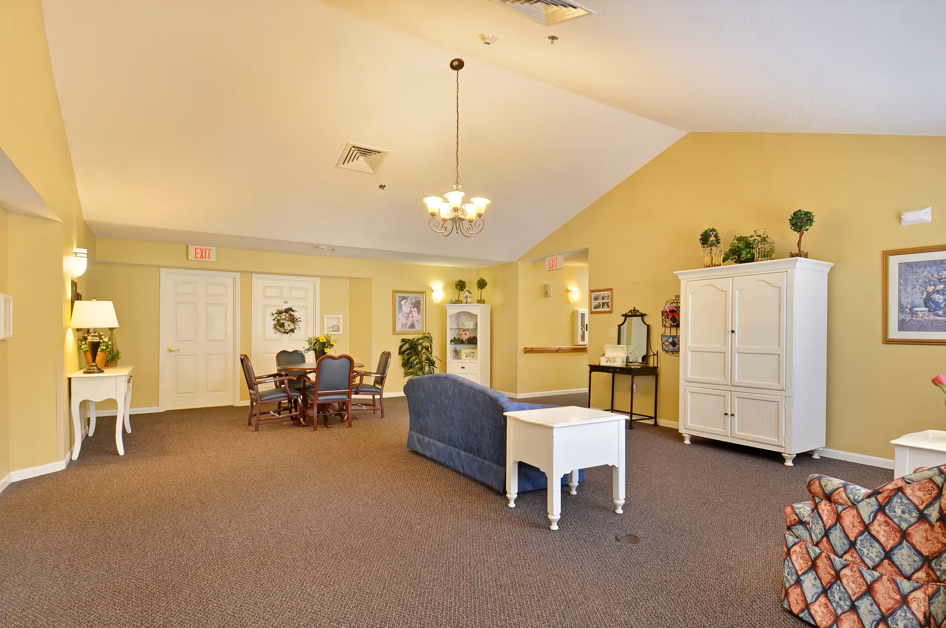 A spacious senior living common area with beige walls and carpeted floor. The room features a blue couch, a white side table, a white cabinet with plants on top, a dining table with chairs, and various decorative plants and framed artwork on the walls. Ceiling lights and wall sconces provide warm lighting.