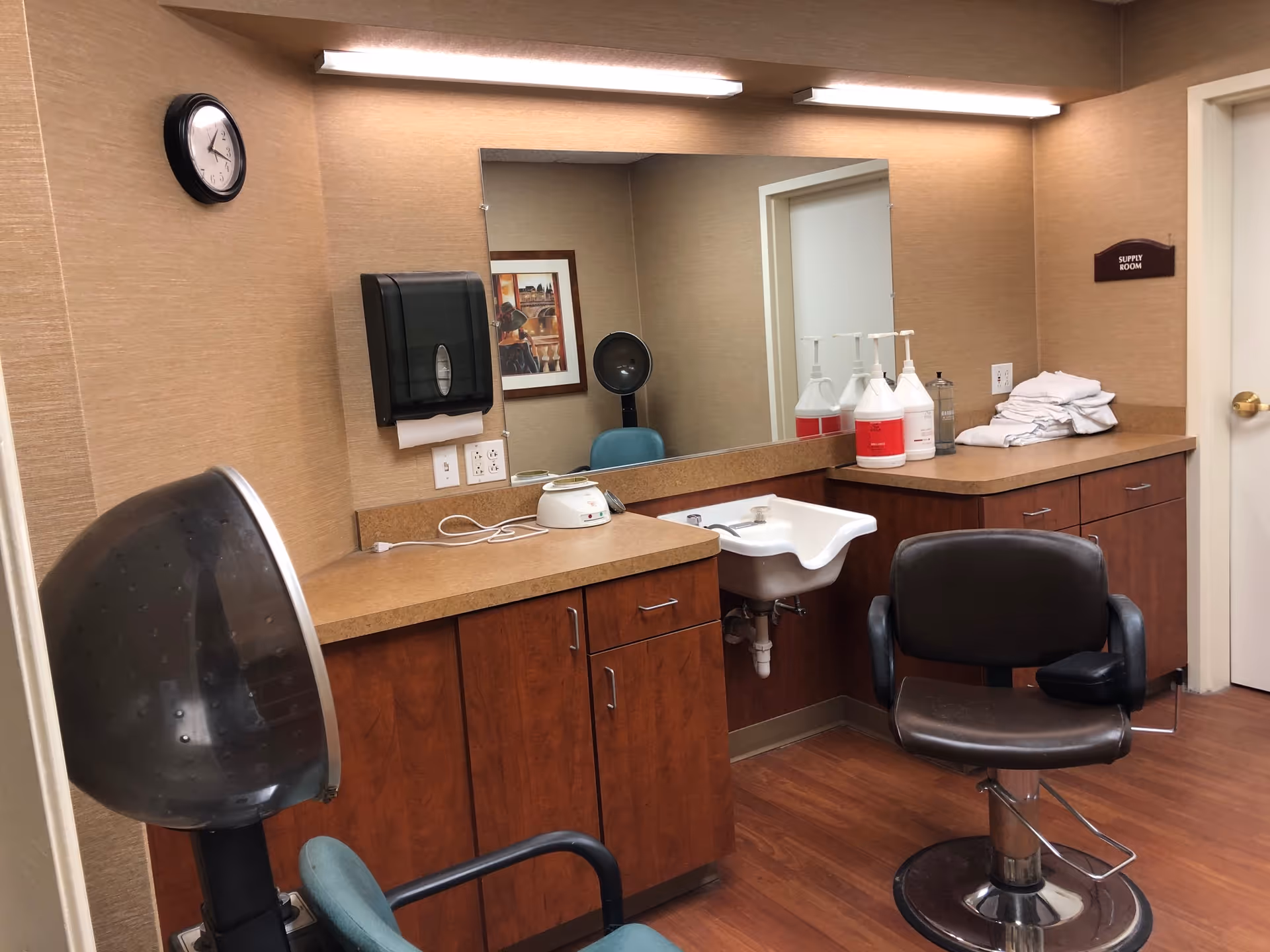 Interior view of a salon or grooming room in a senior living facility with a hair dryer chair, a black salon chair, a sink, countertop with large bottles of hair products, folded towels, a wall clock, and a large mirror reflecting part of the room.
