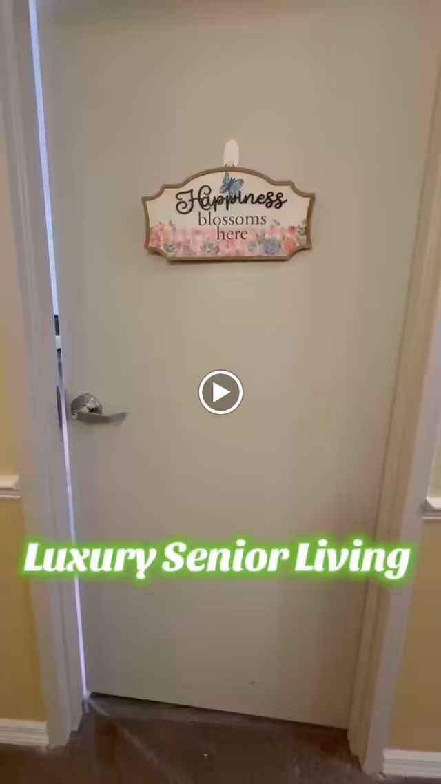 A closed white door with a silver handle in an indoor hallway. On the door hangs a decorative sign with floral designs and a butterfly that reads 'Happiness blossoms here.' The walls around the door are painted light yellow with white trim. Text overlay at the bottom of the image reads 'Luxury Senior Living' in bright green.