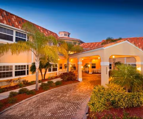 Exterior view of Heron House Assisted Living with a covered walkway, palm trees, and warm lighting.