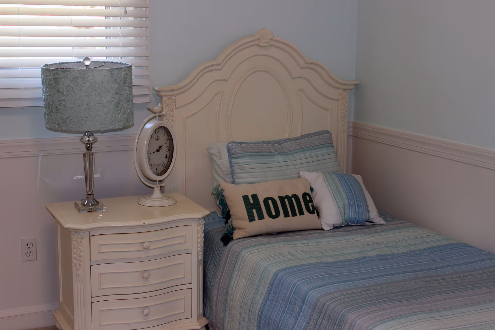 A neatly made single bed with a decorative cream-colored headboard and blue striped bedding. There are three pillows on the bed, including one with the word 'Home' printed on it. Next to the bed is a cream-colored nightstand with three drawers, a decorative table lamp with a blue patterned lampshade, and a round clock. The room has light blue walls with white wainscoting and a window with white blinds.