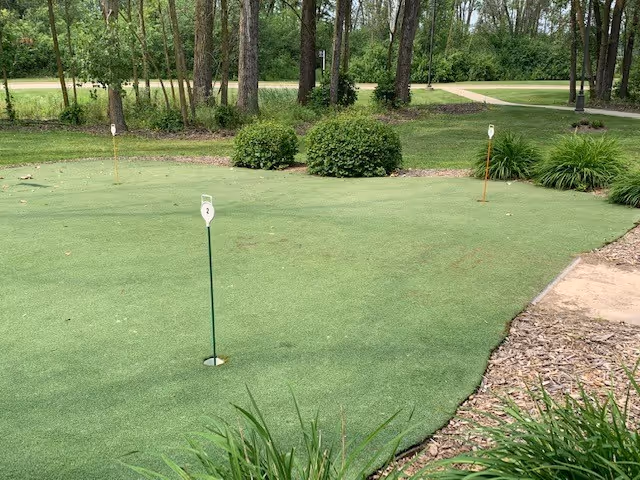 A small outdoor putting green with three holes marked by numbered flags, surrounded by green bushes, trees, and a walking path in the background.