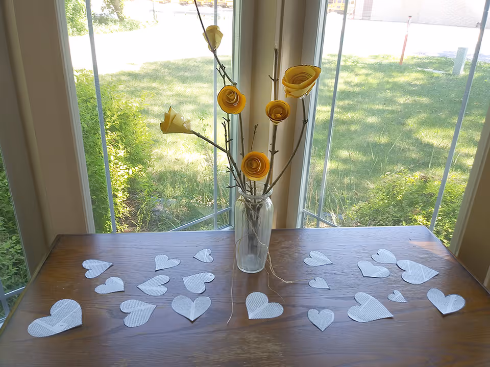 A wooden table in front of a large window with a clear glass vase holding branches with yellow paper flowers. The table is decorated with multiple cut-out paper hearts scattered across its surface. Outside the window, green grass and bushes are visible.