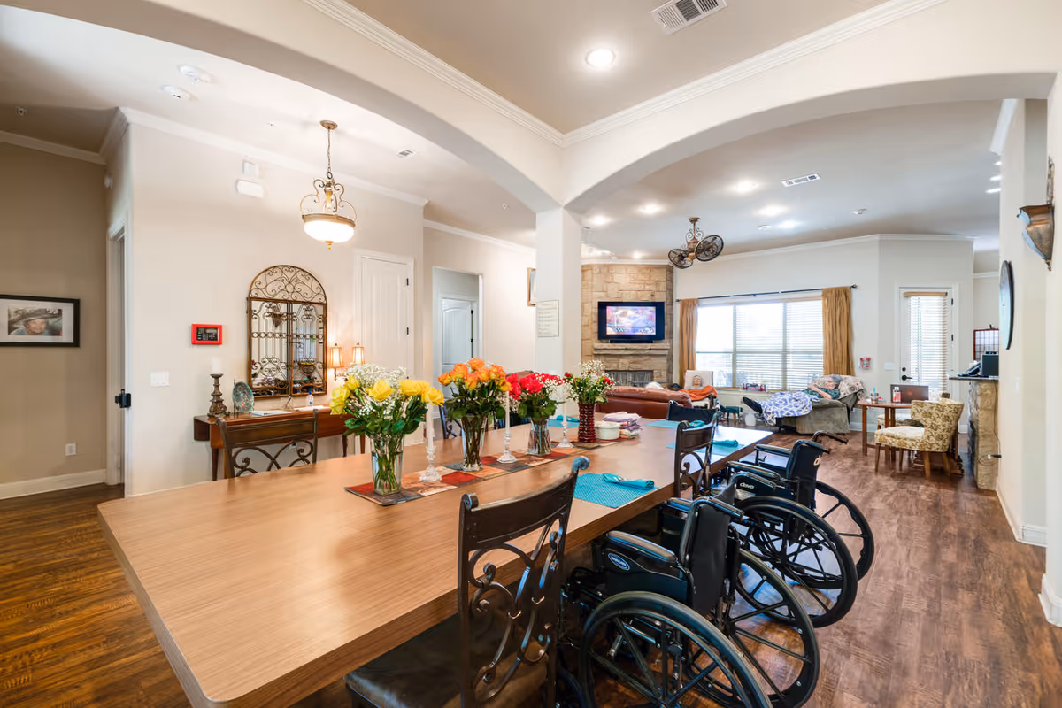 A spacious and well-lit common area in an assisted living facility featuring a large wooden table with floral centerpieces and place settings. Several wheelchairs are positioned around the table. In the background, there is a cozy living room area with a stone fireplace, a mounted TV, comfortable chairs, and large windows with curtains allowing natural light to fill the room.