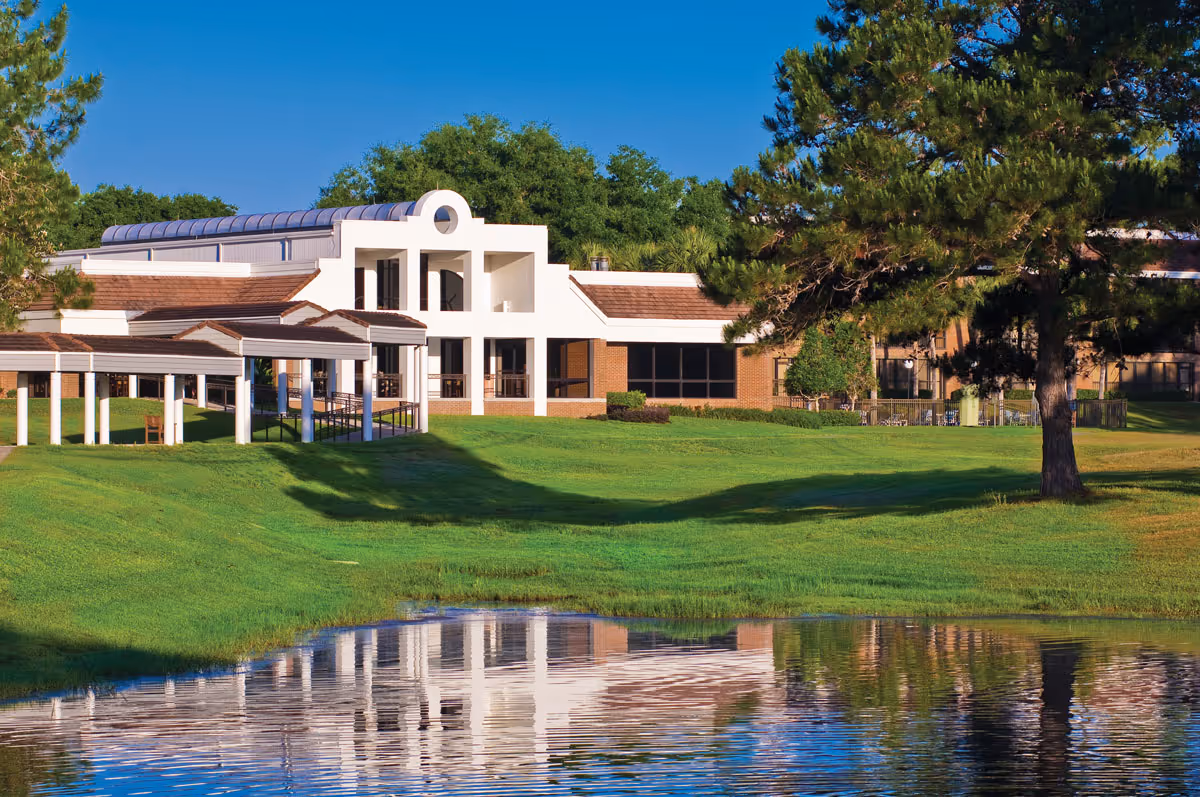 A senior living facility building named Village on the Green with white and brick exterior, surrounded by green grass, trees, and a pond reflecting the building and landscape under a clear blue sky.