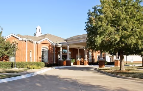 Exterior view of a brick building with a covered entrance supported by white columns, surrounded by trees and landscaping under a clear sky.
