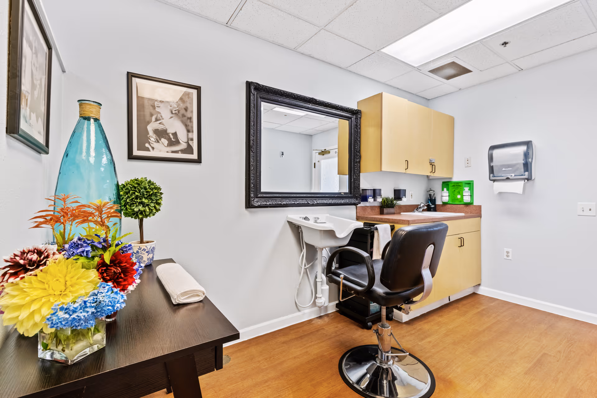 A small salon or grooming area with a black salon chair in front of a white sink. There is a large black-framed mirror on the wall above the sink, wooden cabinets, a paper towel dispenser, and a table with a colorful flower arrangement and a large blue vase. The walls are white and the floor is wooden.