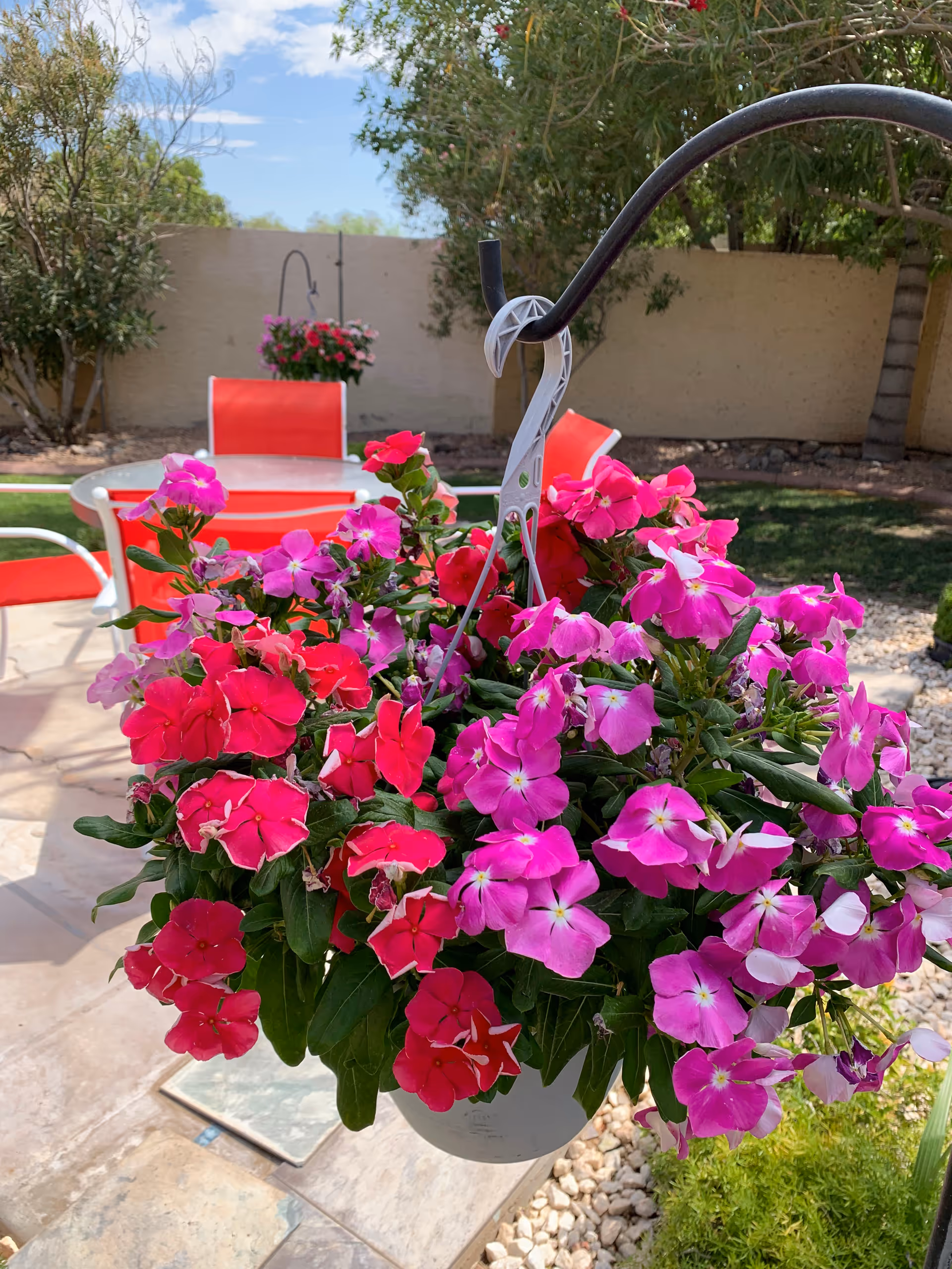A hanging white pot filled with vibrant pink and red flowers in an outdoor garden area. In the background, there are red chairs and a table on a stone patio, with a beige wall and green trees and bushes under a blue sky.