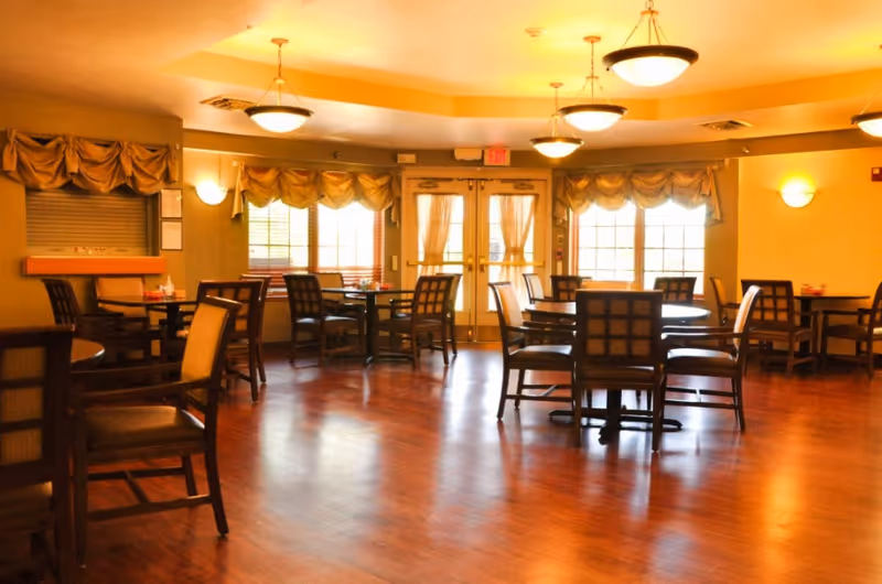 Communal dining room with round tables and wooden chairs on a polished hardwood floor, lit by ceiling fixtures and daylight from curtained windows.