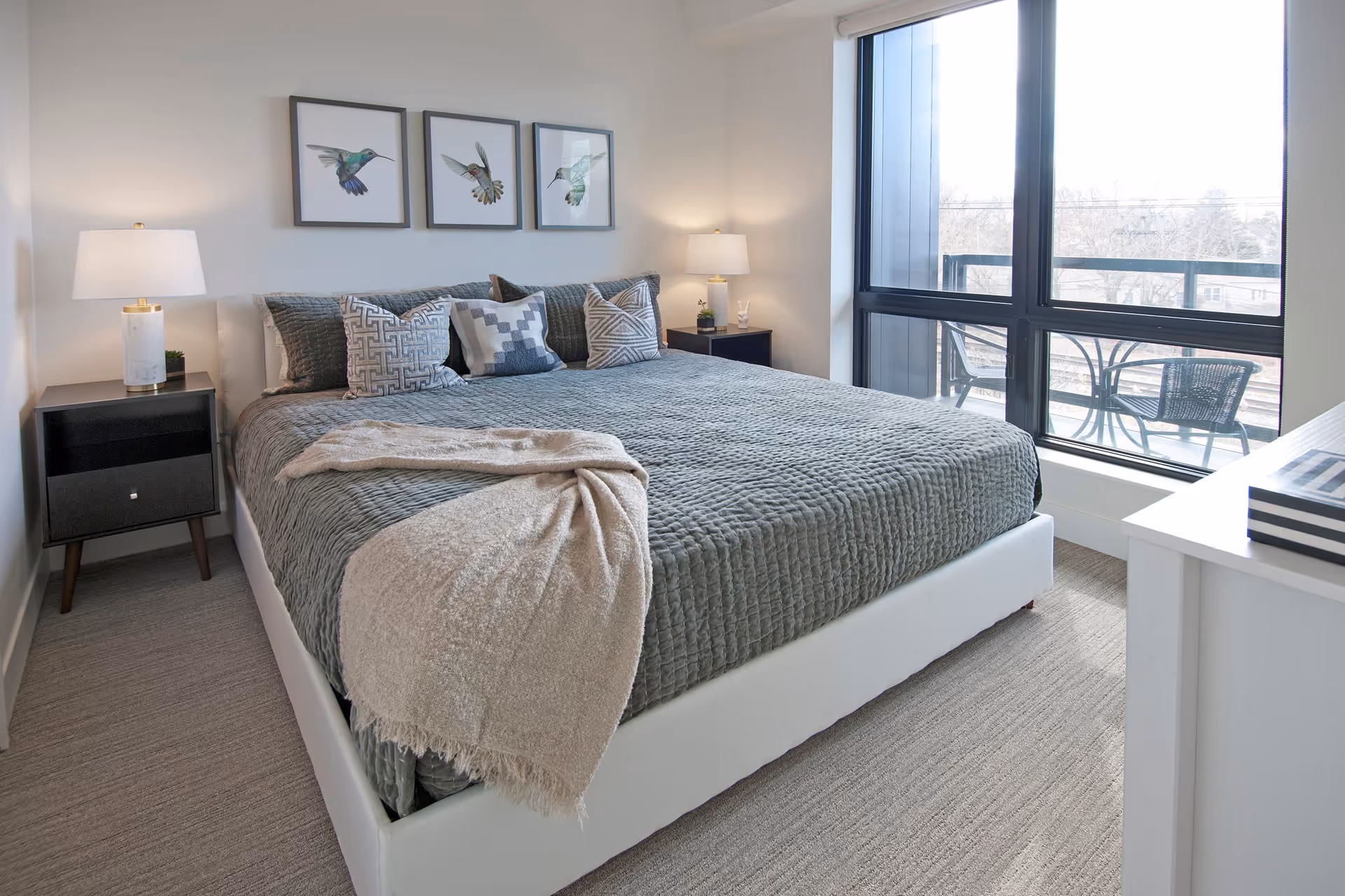 A modern bedroom with a large bed covered in a gray quilt and decorative pillows. Two bedside tables with lamps flank the bed. Three framed pictures of hummingbirds hang on the wall above the bed. A large window reveals an outdoor balcony with a table and chairs.