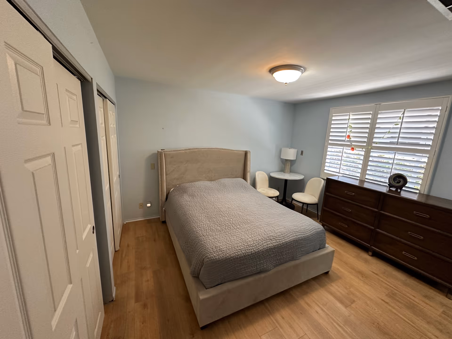 A tidy bedroom with a bed centered, a dresser along the window, two chairs and a round side table beside plantation shutters.