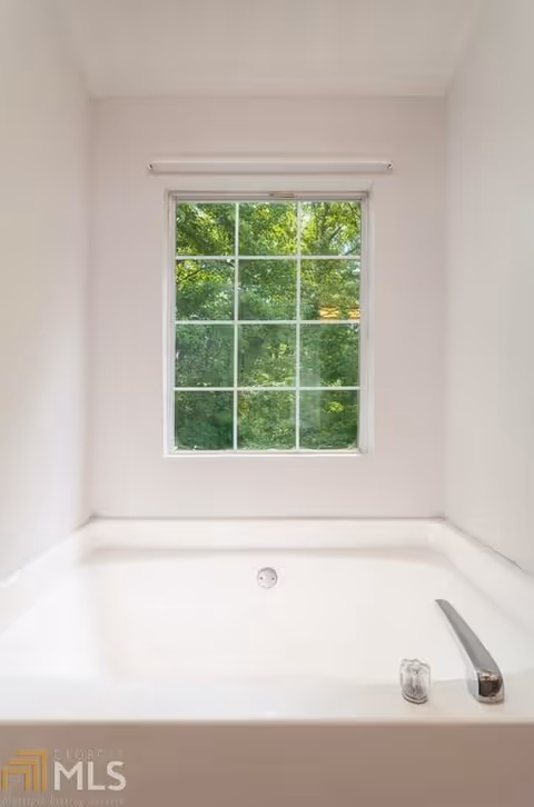 A white bathtub positioned beneath a window with multiple panes showing green trees outside. The bathtub has a silver faucet and a clear knob on the right side.