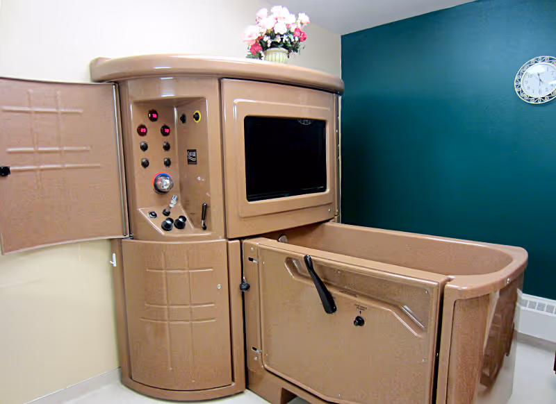 A large accessible walk-in bathing tub with control panel in a room with a teal accent wall, clock, and a vase of flowers on top.