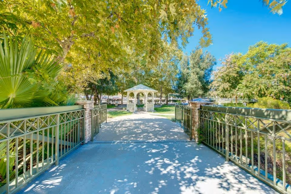 A sunny outdoor pathway leading to a white gazebo surrounded by green trees and plants, with metal railings on both sides of the path.