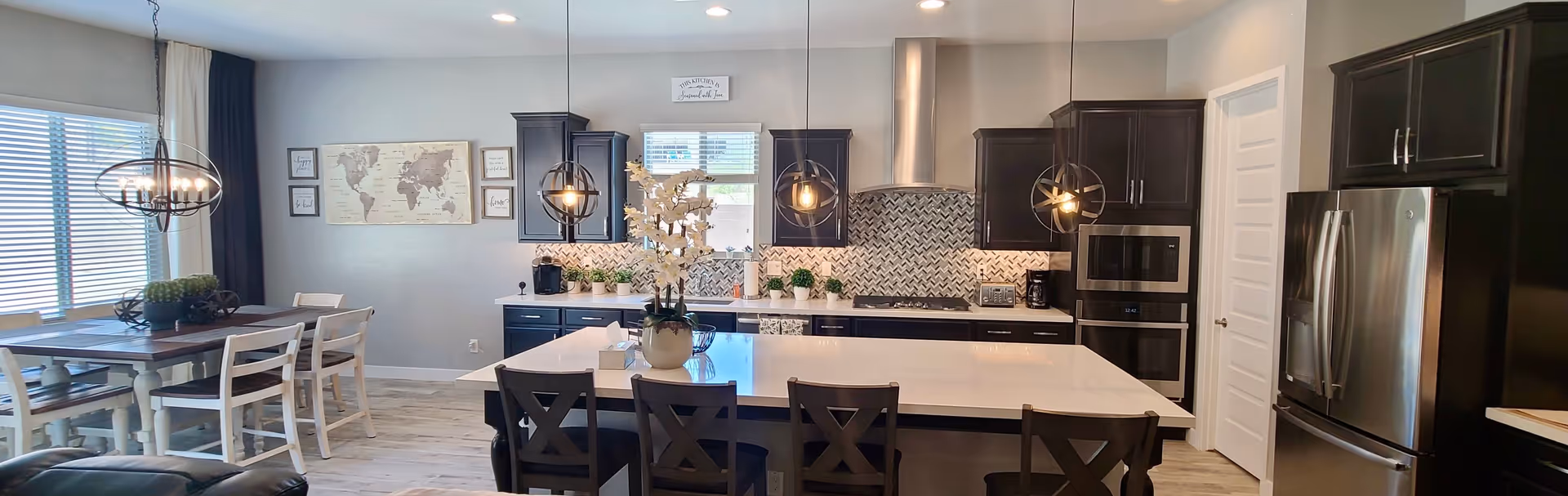 A modern kitchen and dining area featuring a large white island with four dark wooden chairs, three pendant lights hanging above, dark cabinetry, stainless steel appliances including a refrigerator and built-in oven, a patterned backsplash, and a dining table with white chairs near a window with blinds.
