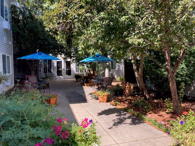 Outdoor courtyard area with concrete pathways, garden beds with flowers and greenery, several trees providing shade, and patio tables with blue umbrellas and chairs. The area is adjacent to a building with windows and doors.
