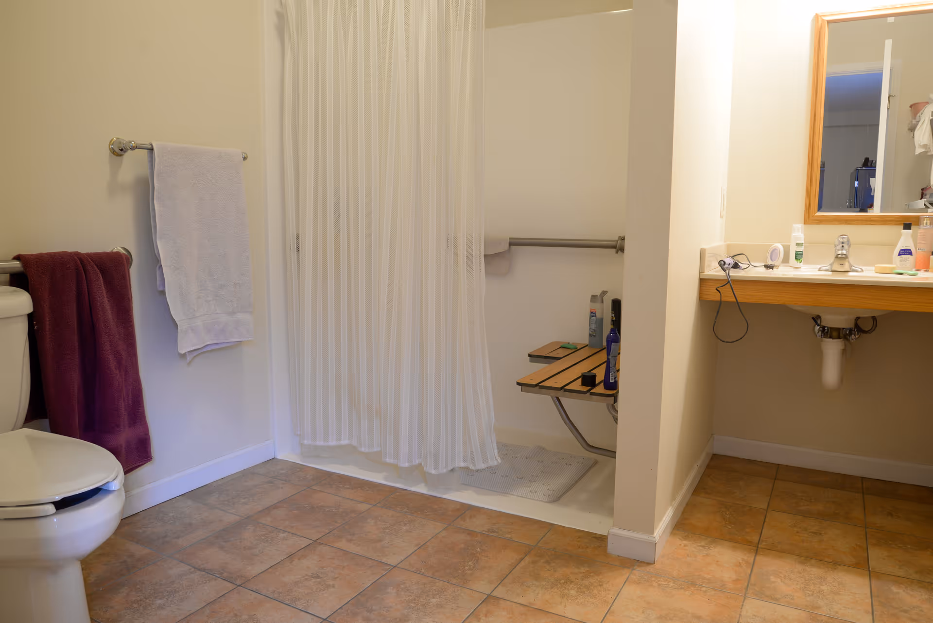 A bathroom with a toilet on the left side, two towels hanging on towel racks, a walk-in shower with a white curtain and a wooden bench inside, and a sink with a mirror above it on the right side. Various toiletries are placed on the sink counter.