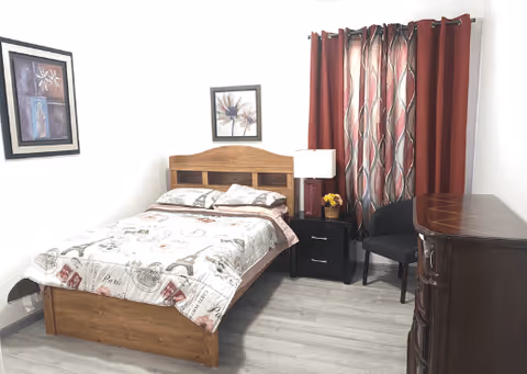 A bedroom with a wooden bed frame and a bed made with a patterned comforter. There are two framed floral pictures on the white walls. Next to the bed is a black nightstand with a lamp and a small flower arrangement. A dark chair is placed near the nightstand, and red and beige patterned curtains cover the window. A dark wooden dresser is visible on the right side of the image. The floor is light-colored wood.