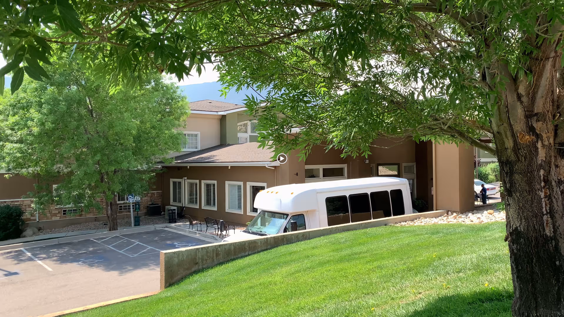 Exterior view of a senior living facility with a beige building partially visible behind green trees and a grassy hill. A white shuttle bus is parked near the building, and there is a small parking area with marked spaces. The scene is shaded by tree branches overhead.