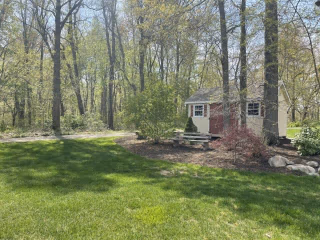 A small beige and red garden shed surrounded by trees and bushes in a grassy outdoor area with a dirt path nearby.