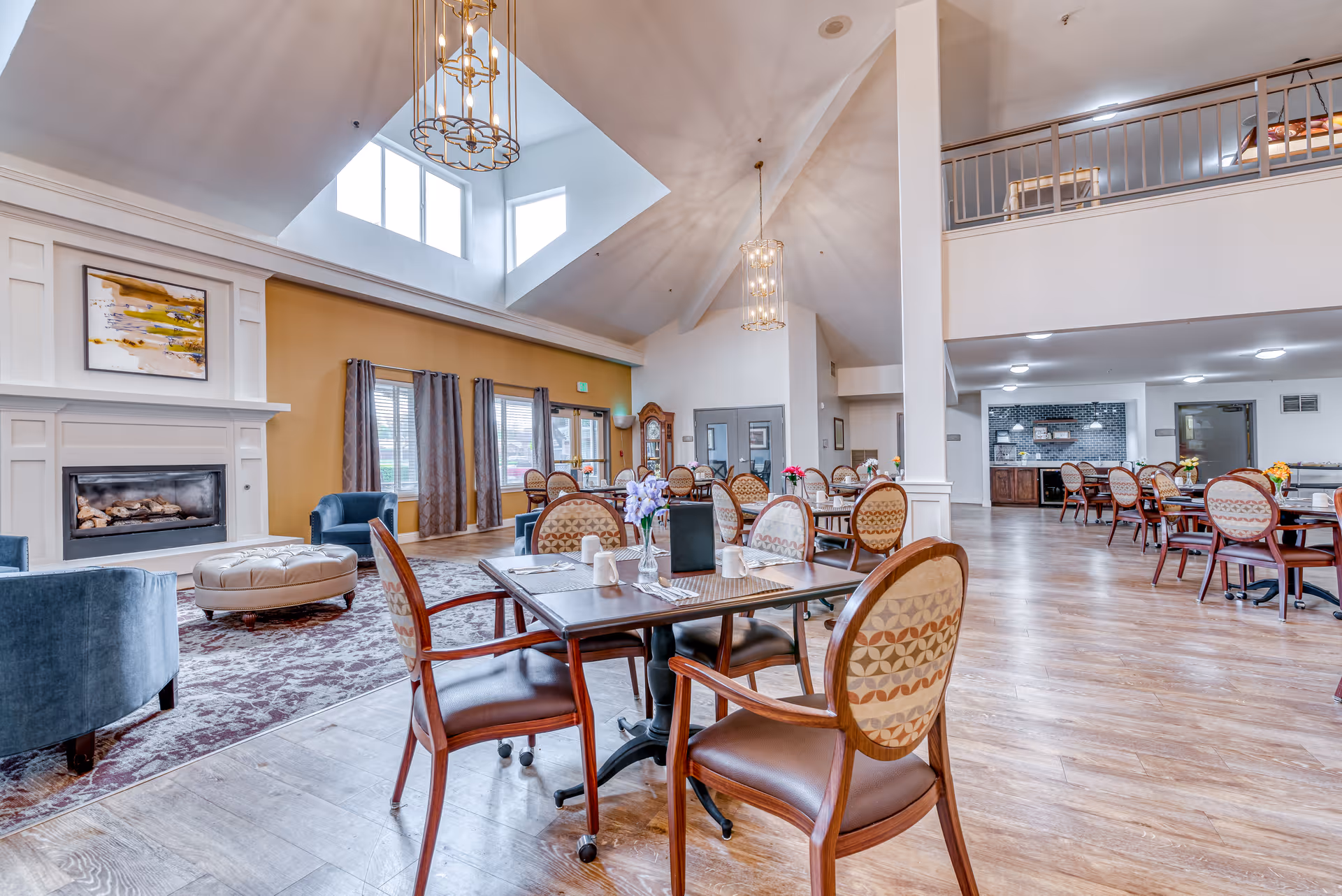 Spacious senior living communal dining and lounge area with tables and chairs, a fireplace, and a high vaulted ceiling with skylights.