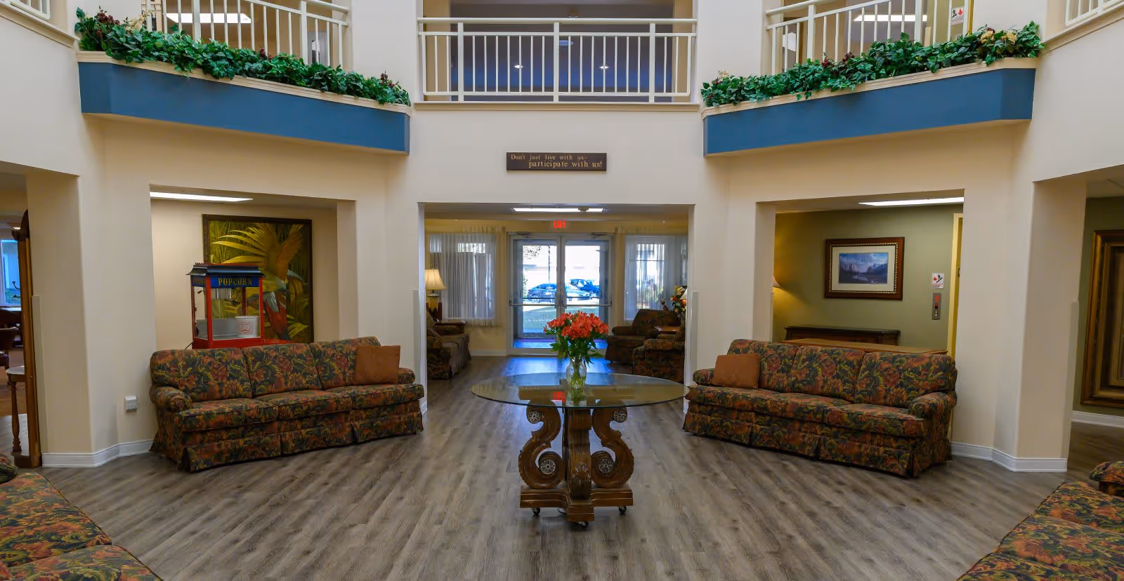 A spacious common area with floral patterned sofas arranged around a central glass-top table with a wooden base. The table holds a vase with red flowers. The room has light-colored walls and wood flooring. Above the entrance door, there is a sign that reads 'Don't just live with us- participate with us!'. There are plants on ledges above the sofas and framed artwork on the walls.