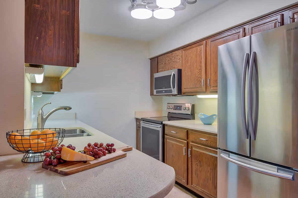 A modern kitchen with wooden cabinets, a stainless steel refrigerator, microwave, and stove. The countertop features a wire basket with oranges and a wooden cutting board with grapes and a wedge of cheese. The kitchen is well-lit with ceiling lights and under-cabinet lighting.