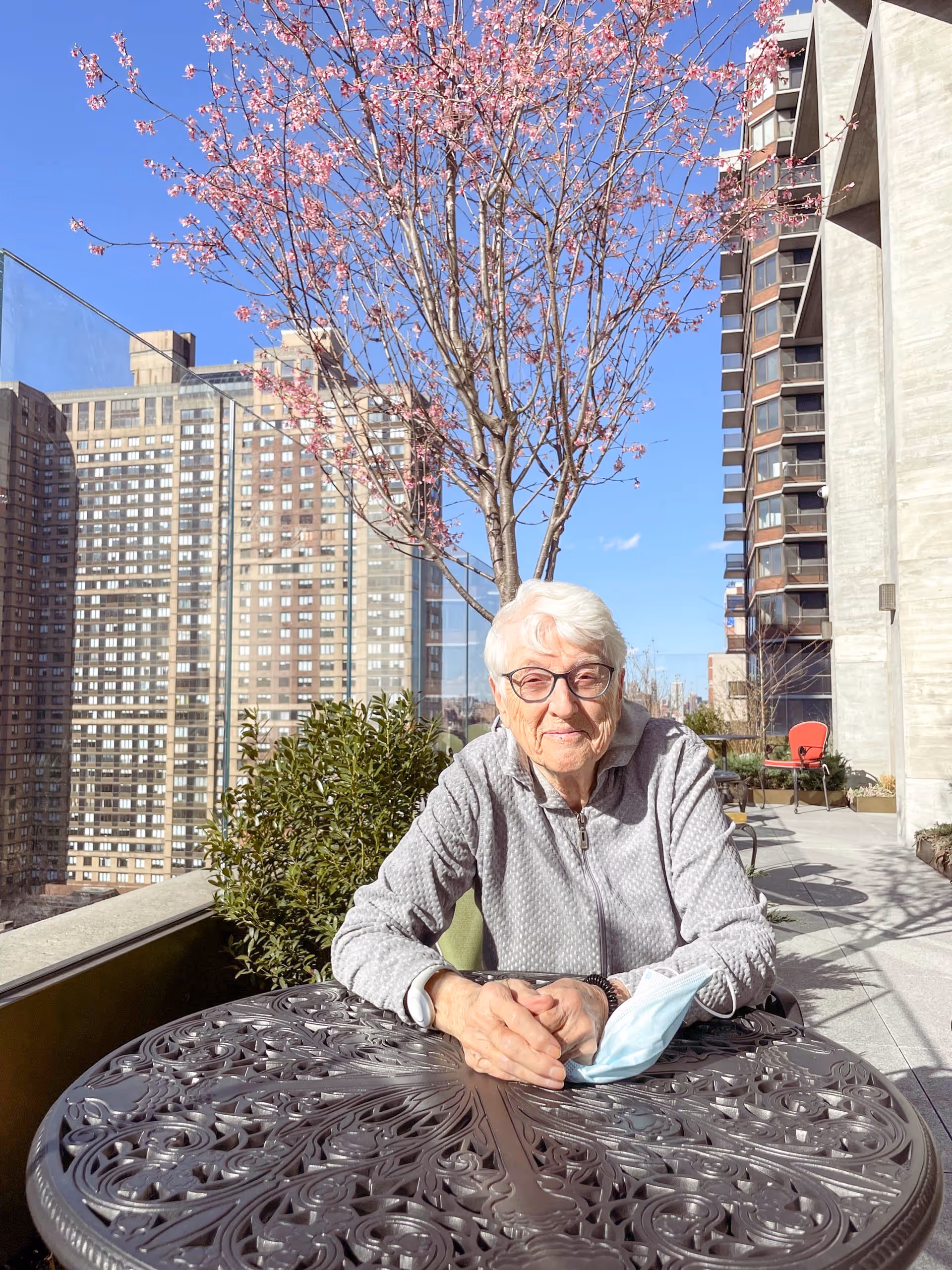 An elderly woman with white hair and glasses sits at an ornate metal outdoor table on a balcony or terrace. She is wearing a light gray zip-up jacket and has a light blue face mask resting on the table in front of her. Behind her, there is a blooming tree with pink flowers, green shrubbery, and tall apartment buildings under a clear blue sky.