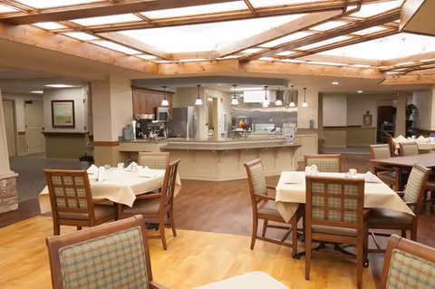 A dining area in a senior living facility with several tables covered with beige tablecloths and set with white napkins and utensils. The room features wooden chairs with plaid upholstery, a wooden floor, and a ceiling with a large skylight. In the background, there is a kitchen area with stainless steel appliances and pendant lighting.