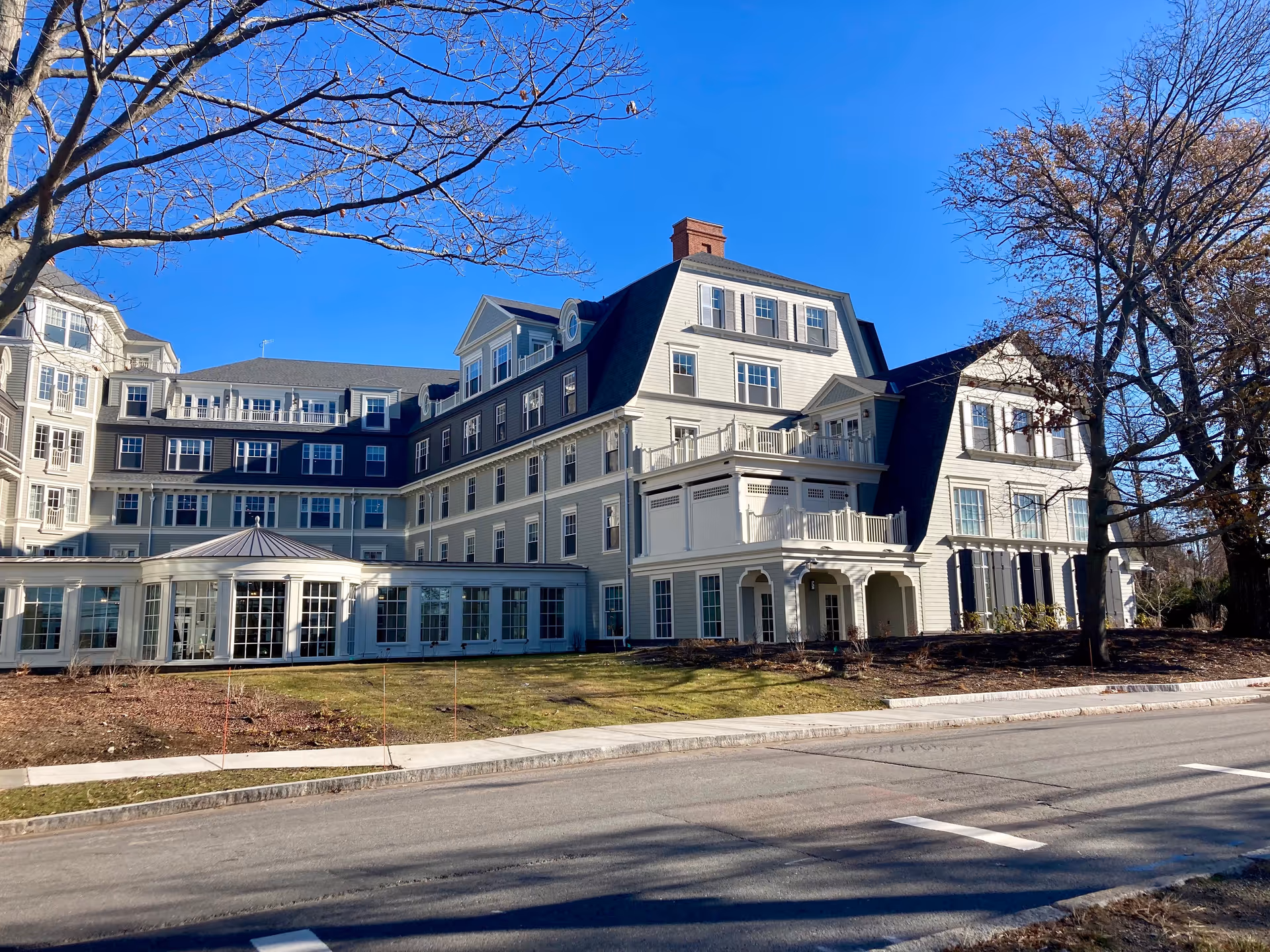 Multi-story light-colored residential building with many windows, balconies and a porch facing a street under a clear blue sky.