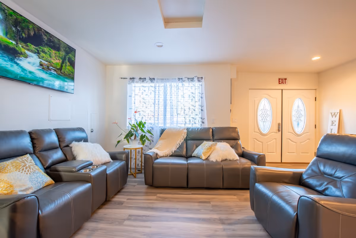 A bright living room with three dark gray leather sofas arranged around a window with sheer floral curtains. The sofas have decorative pillows and a throw blanket. There is a small round side table with a plant on it next to the window. A large flat-screen TV mounted on the wall displays a nature scene with a river and greenery. Double white doors with oval glass panels and an exit sign above them are visible in the background. The room has light wood flooring and recessed lighting.