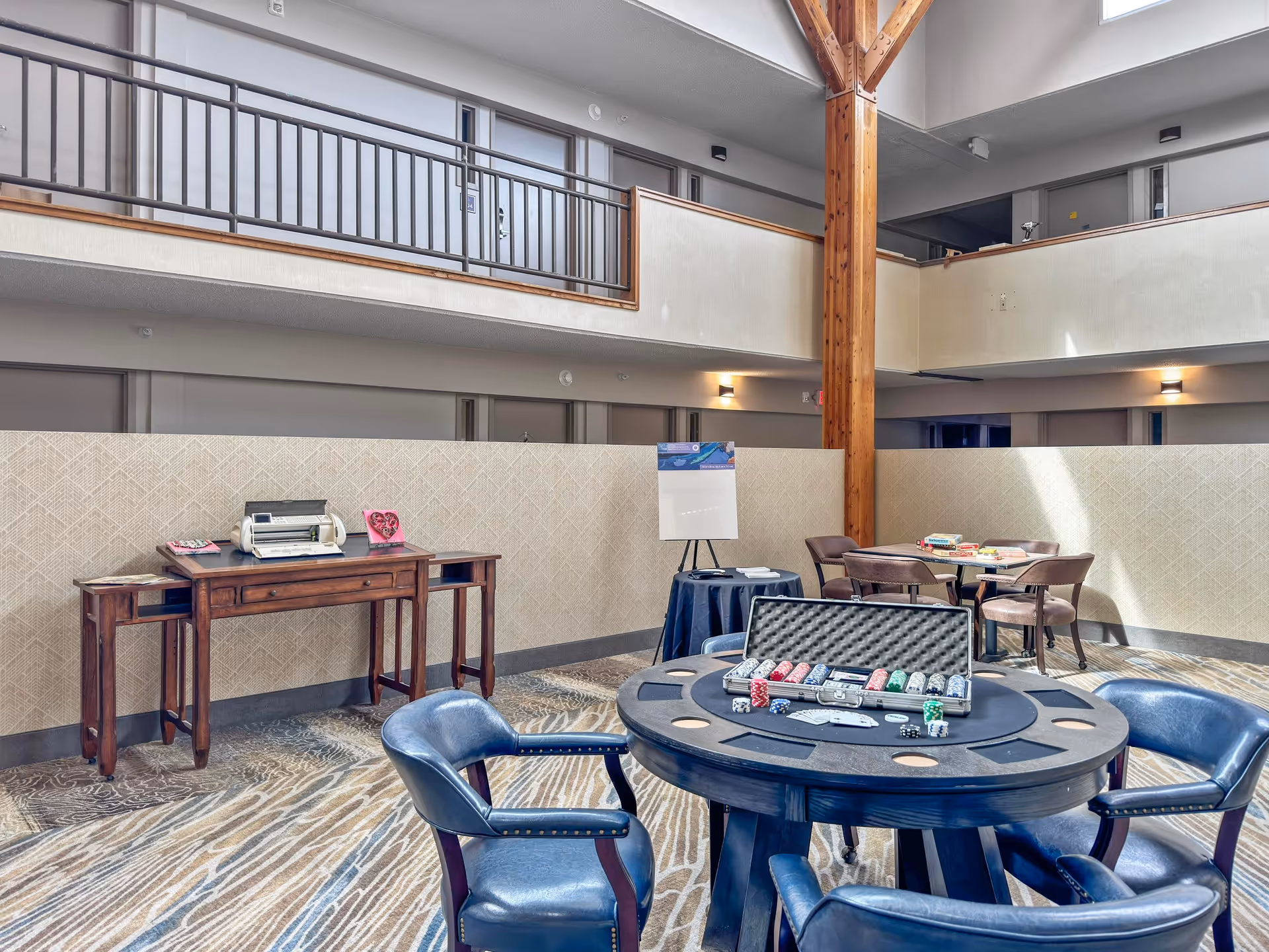 A communal indoor space with a poker table in the foreground featuring poker chips and cards, surrounded by blue leather chairs. In the background, there is a wooden table with a crafting machine and some decorations, and another table with board games and chairs. The area has a high ceiling with wooden beams and an upper balcony with railings.