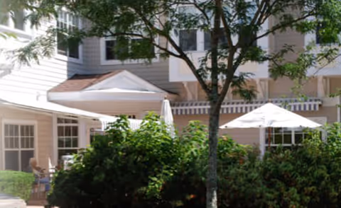 Outdoor patio area at Tamarisk Senior Living with white umbrellas, chairs, and tables surrounded by greenery and trees in front of a multi-story building.